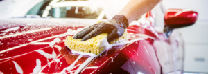 Arm und Hand mit Handschuh wäscht ein rotes Auto mit einem gelbem Schwamm.